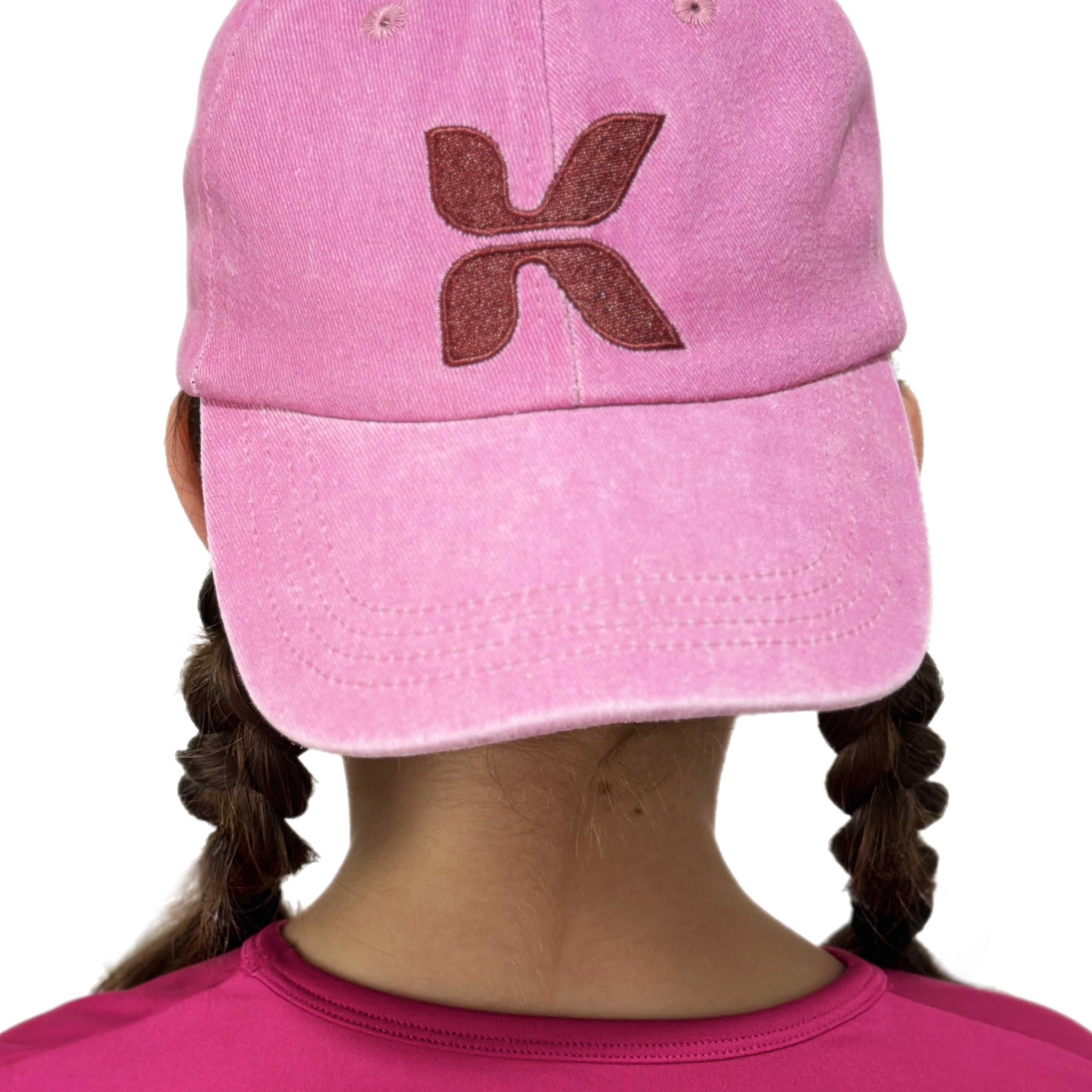 Pink cap with a brown logo worn by a tween girl, with a matching pink shirt underneath on a white background.
